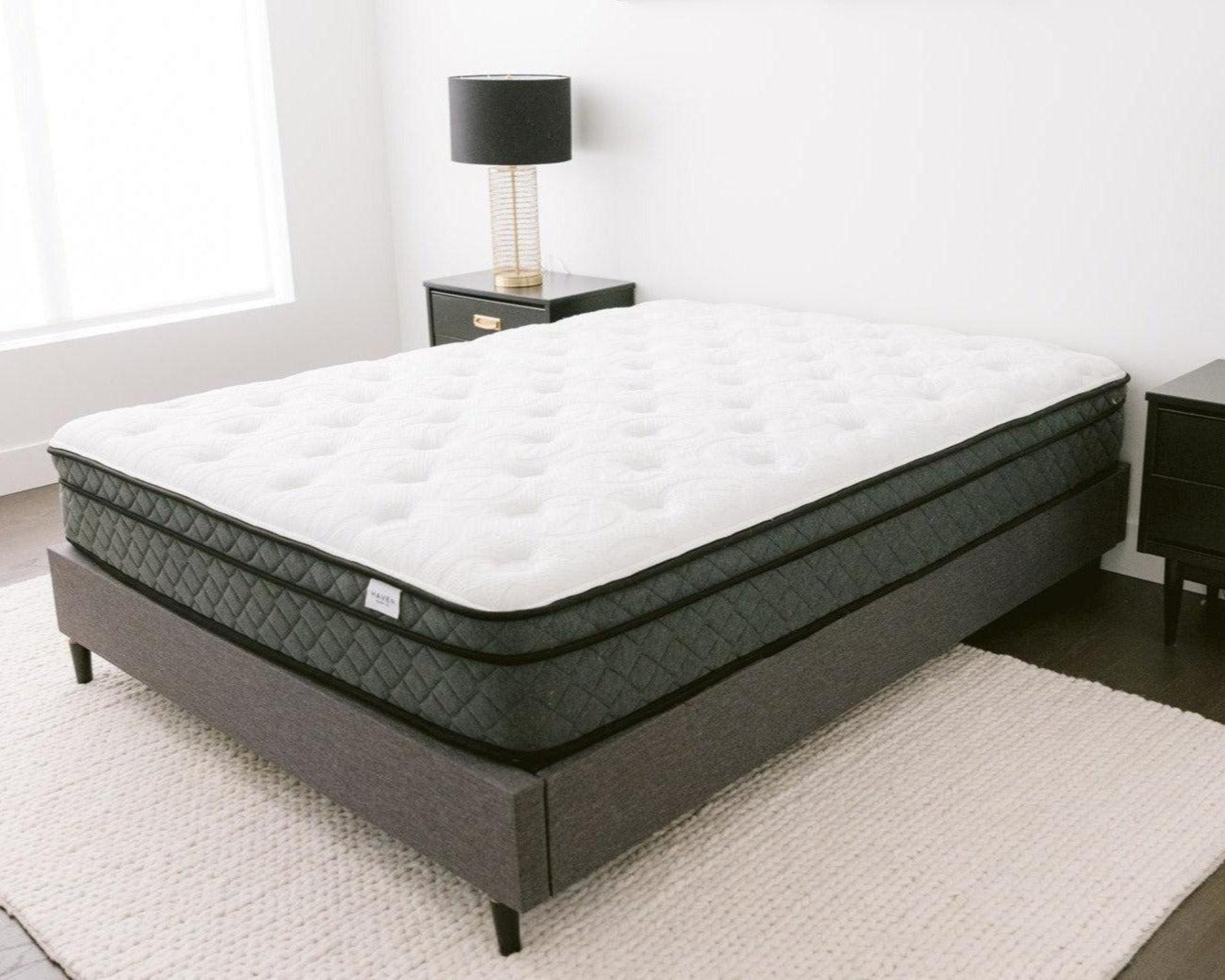 Image of a Haven Pocket Coil Mattress with a white quilted top and dark grey sides, displayed in a modern bedroom setting with a grey upholstered bed frame, black nightstands, and a light-colored area rug. The mattress features 884 individually wrapped coils, a bamboo cover with cool touch technology, and plant-based foams for enhanced sleep comfort and support. Made in Canada.