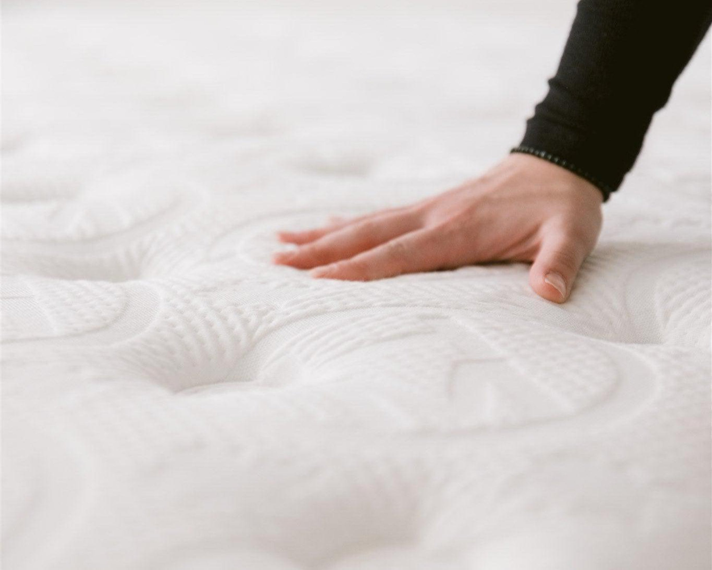 Close-up of a hand pressing down on the white quilted surface of the Haven Pocket Coil Mattress, demonstrating the plush texture and support offered by the mattress's plant-based foams and premium construction. The mattress is designed to provide optimal comfort with 884 individually wrapped coils and a bamboo cover with cool touch technology.