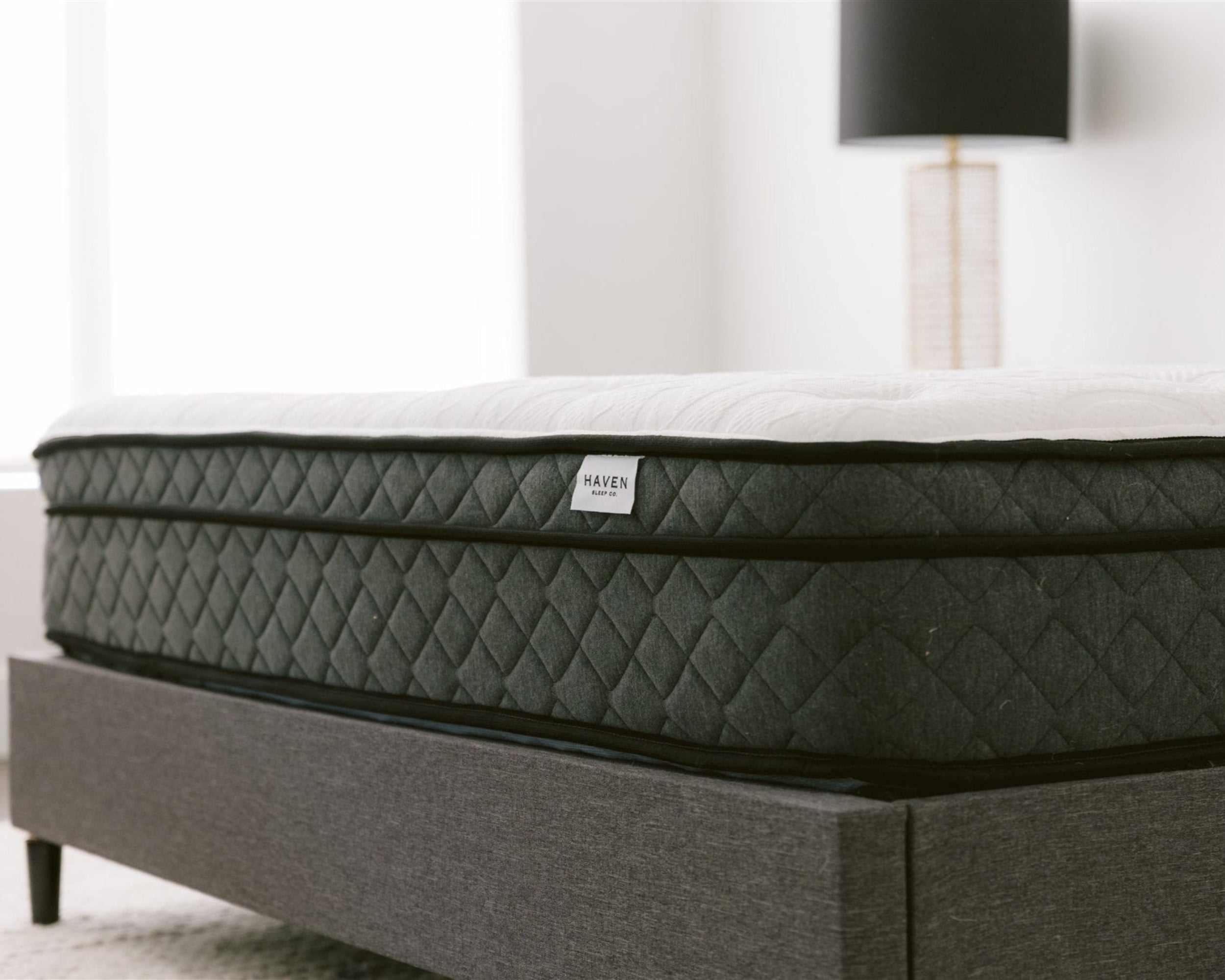 Close-up side view of the Haven Pocket Coil Mattress with a white quilted top and dark grey quilted sides, resting on a grey upholstered bed frame. The mattress, featuring a label with the Haven Sleep Co. logo, showcases its premium construction designed to provide optimal support with 884 individually wrapped coils and a bamboo cover with cool touch technology.