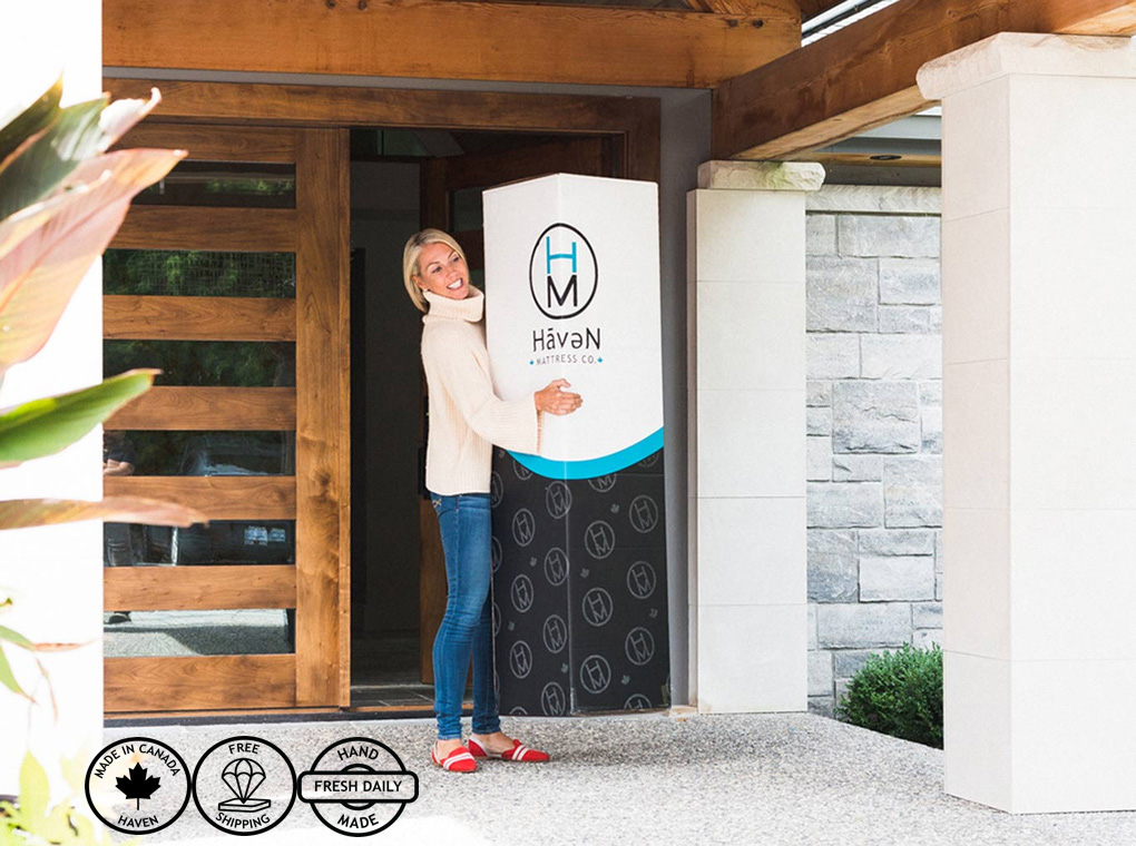 Woman standing at front door smiling and holding a Haven mattress box delivered to her home.