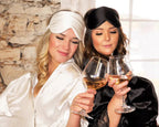 Two women in luxurious satin sleepwear, one in white and the other in black, enjoy a glass of wine while wearing satin eye masks from the Dream Satin Sleep Set. The set includes a satin pillowcase, scrunchie, and eye mask, offering a cruelty-free and sustainable alternative to traditional silk, ensuring frizz-free hair and a rejuvenating sleep.