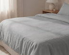 Gray bedding set on a bed in a bedroom with a lamp and nightstand.
