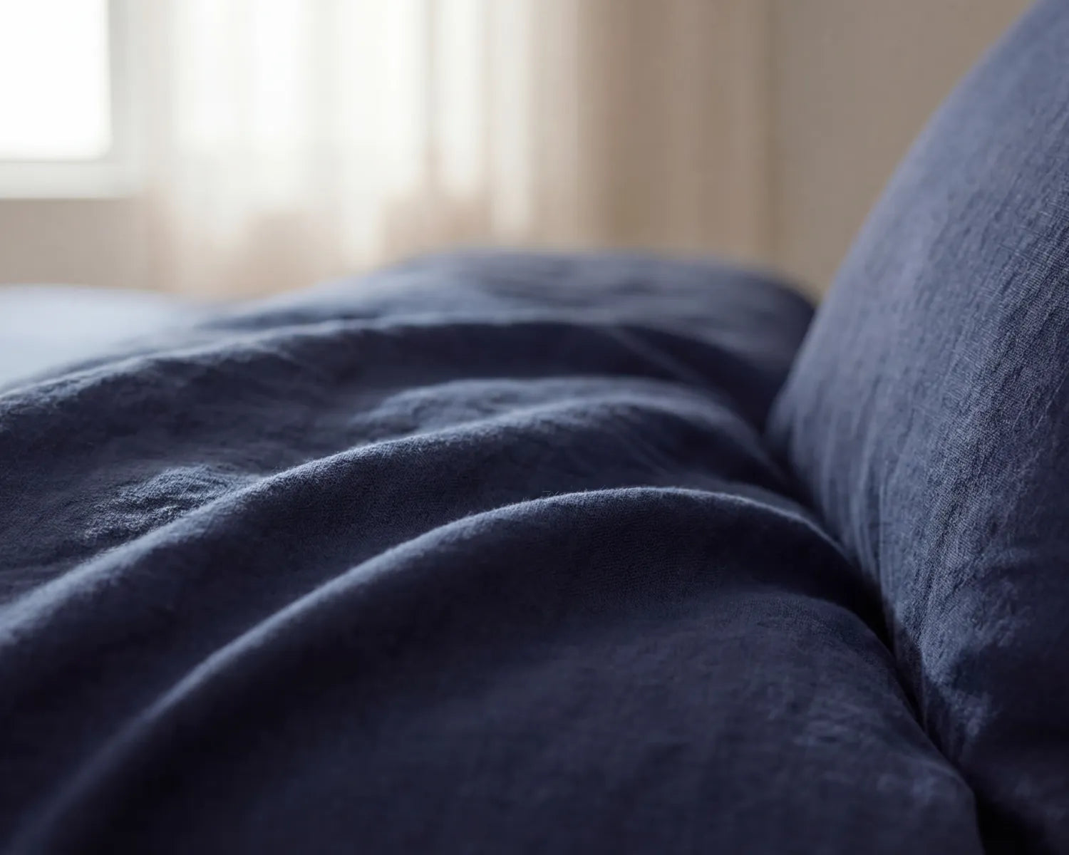 Close-up of a blue textured blanket and pillow on a bed with a blurred window in the background.