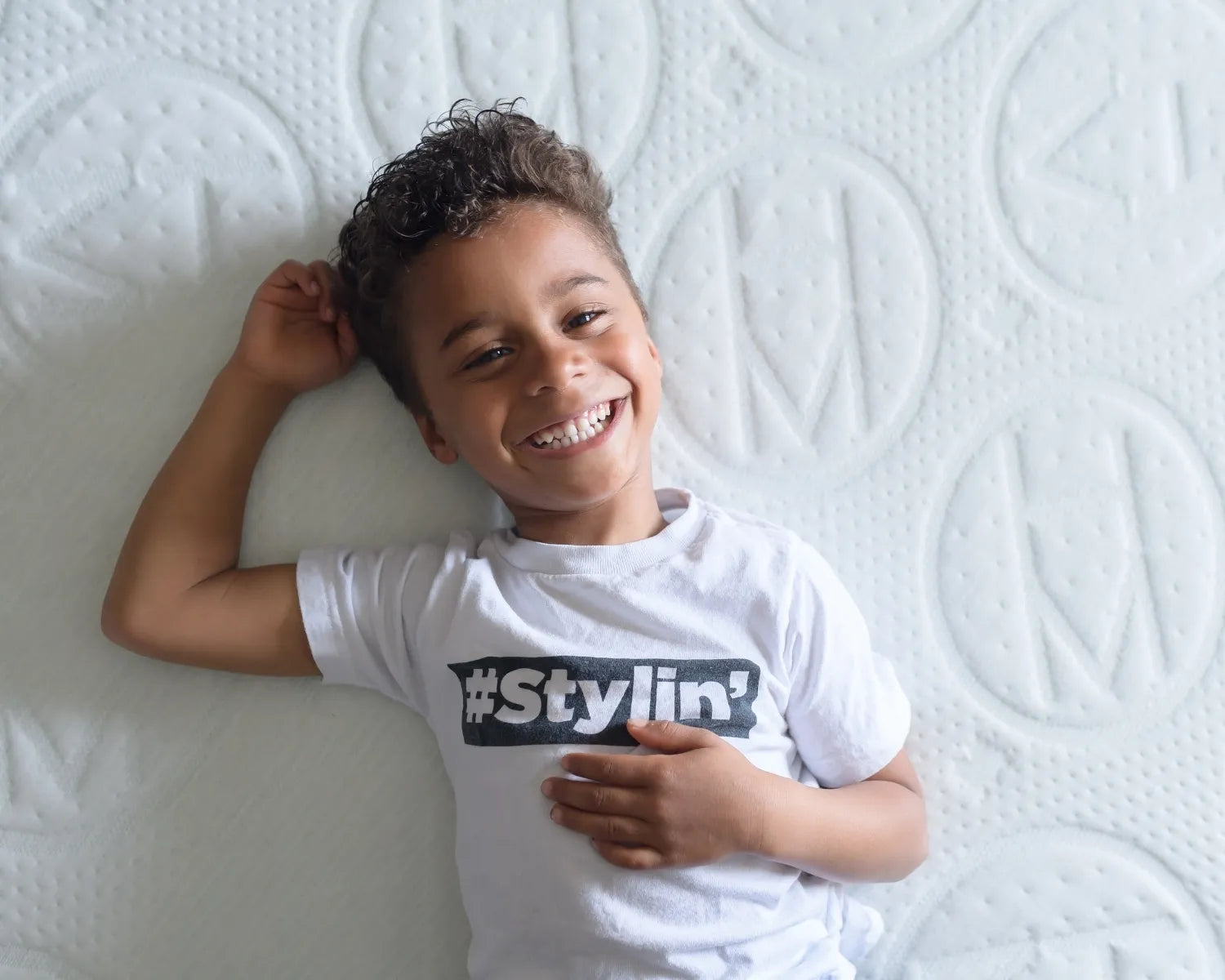 Child wearing a white t-shirt with '#Stylin'' text, lying on a textured white surface.