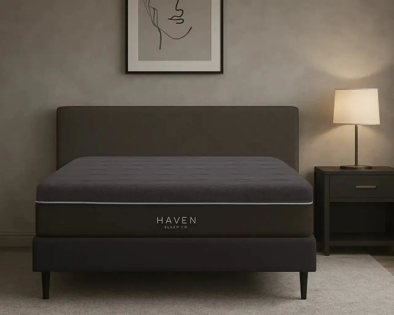 Dark gray Haven mattress in a bedroom setting with a lamp and framed art on the wall.