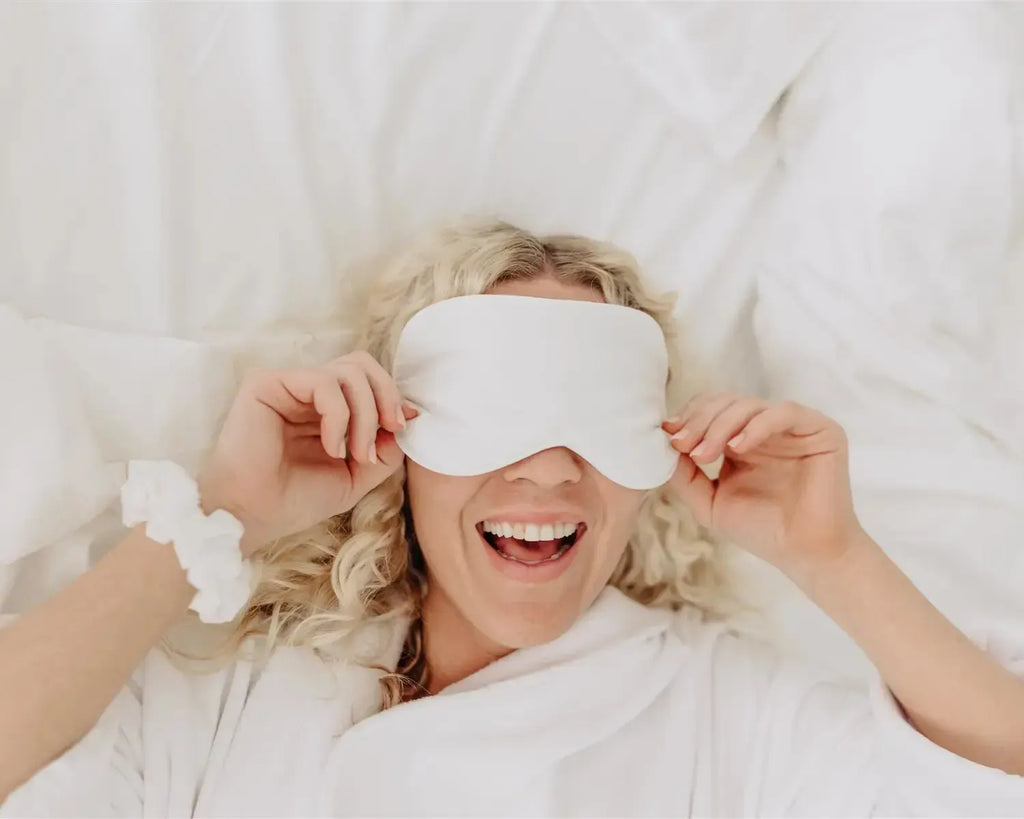 Woman wearing a white sleep mask in bed, smiling.