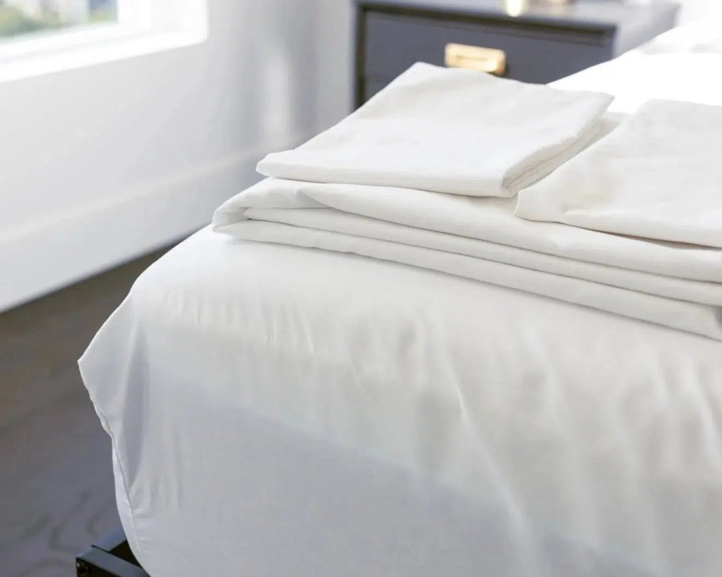 Stack of folded white Bedface Percale sheets on a neatly made bed in a modern bedroom setting with a dark bedside table.