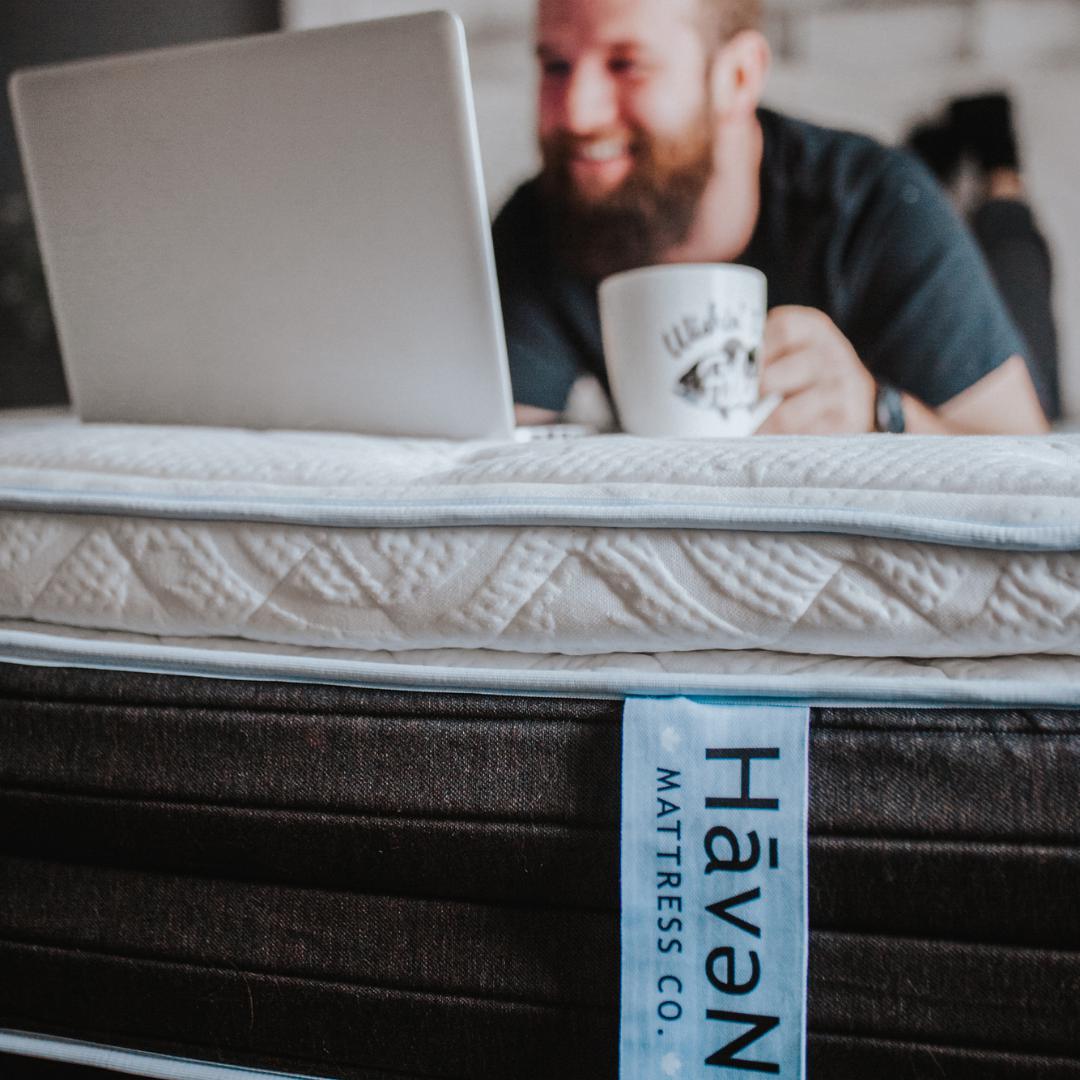 Man lying across Haven Mattress holding a cup of coffee looking at a white laptop with Haven mattress tag down front