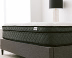 Side view of the Haven Pocket Coil Mattress with a white quilted top and dark grey diamond-patterned sides, positioned on a grey upholstered bed frame in a modern bedroom. The mattress label displays the Haven Sleep Co. logo, emphasizing its premium construction, including 884 individually wrapped coils and a bamboo cover with cool touch technology.