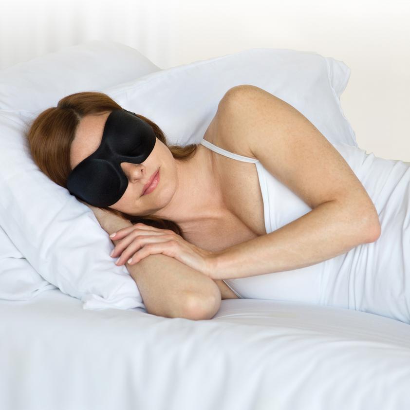 Woman in white with a pair of Bedface Blackout 40 blinks sleep mask in black sitting on head pulling her hair back as she is lying on memory foam pillows on a Haven Mattress tucked into Stralight White bedding