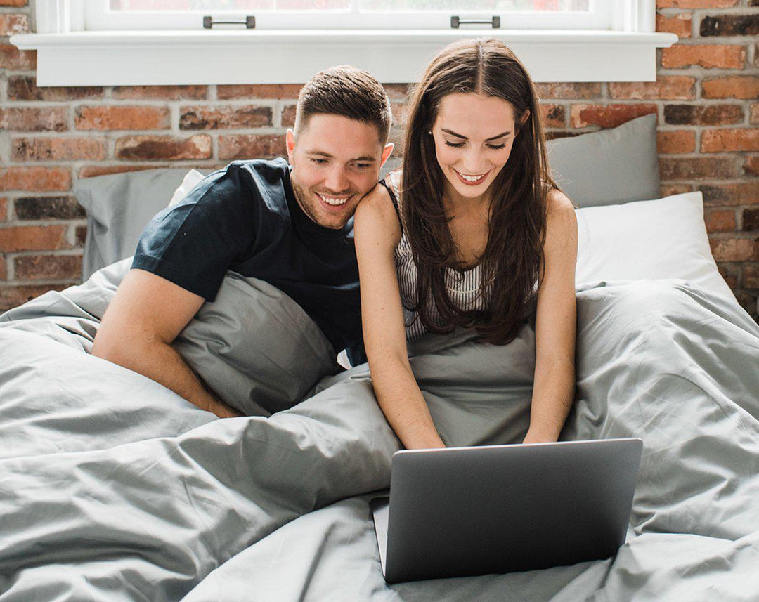 Man and woman cozied up in Haven Mattress bed-in-box bed, tucked into Storm Grey Bedface sheets and duvet cover. The couple is both looking at a grey laptop while the woman is typing 