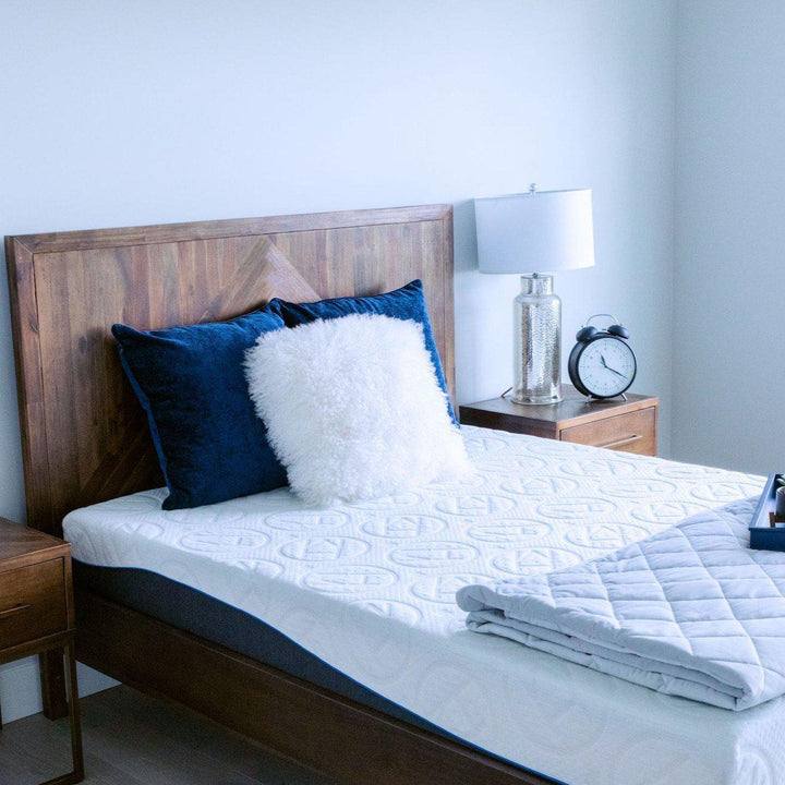 10" Rejuvenate Haven Mattress on a wooden frame with headboard and end tables with a two navy blue throw pillows and a cozy white fuzzy throw pillow at top, and a weighted pastel blue blanket folded up at bottom